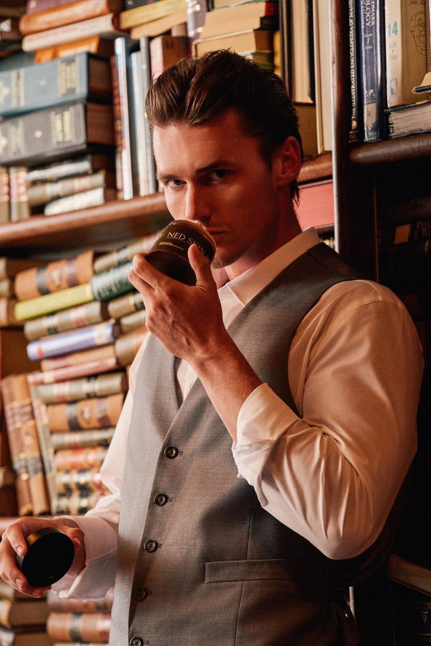 A man in a vest by a bookshelf, lifting his hand to his face