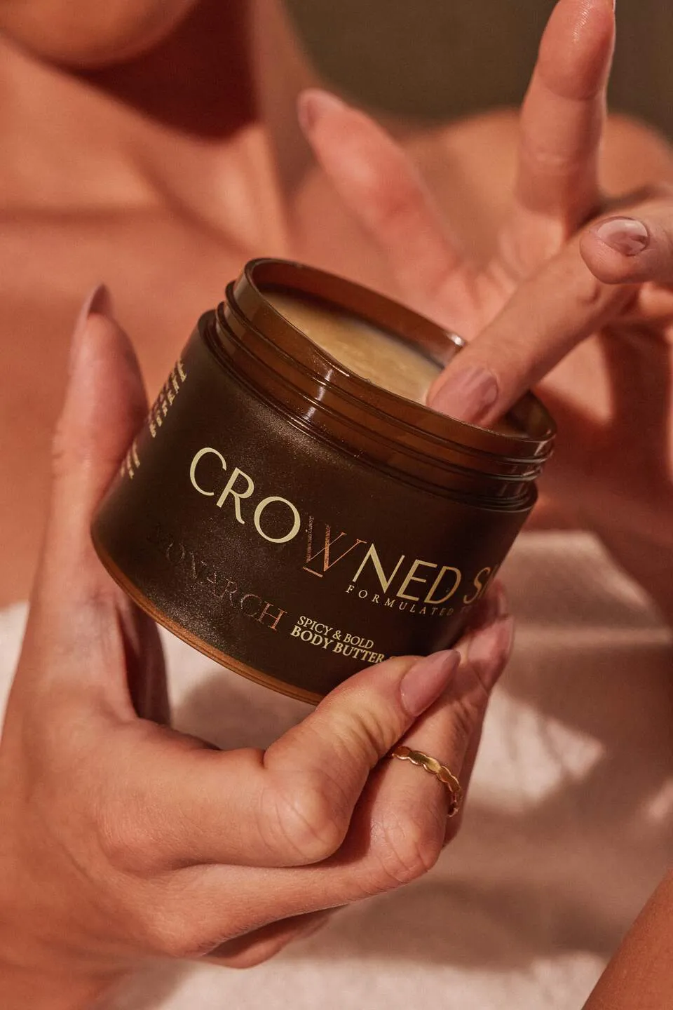 Hands holding a Crowned Skin body butter jar with a gold emblem on the lid