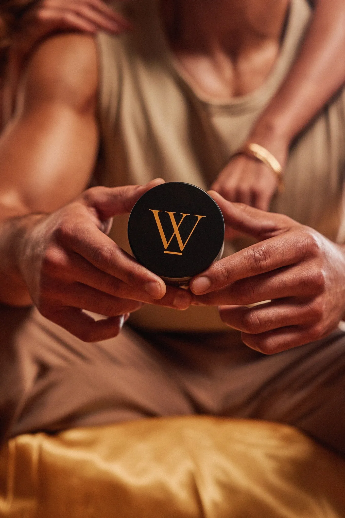 Hands holding an open jar of Crowned Skin body butter against gold fabric